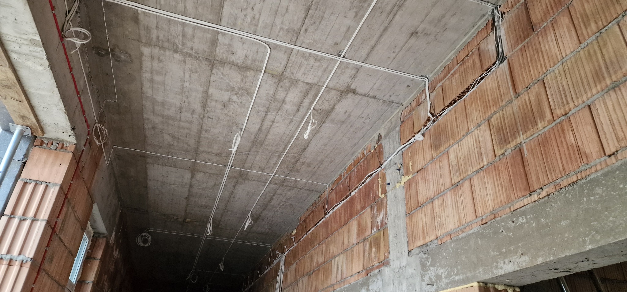Surowy betonowy strop z rozciągniętymi białymi kablami elektrycznymi, ściany z czerwonej cegły z widocznymi przewodami w bruzdach, instalacja w trakcie realizacji.