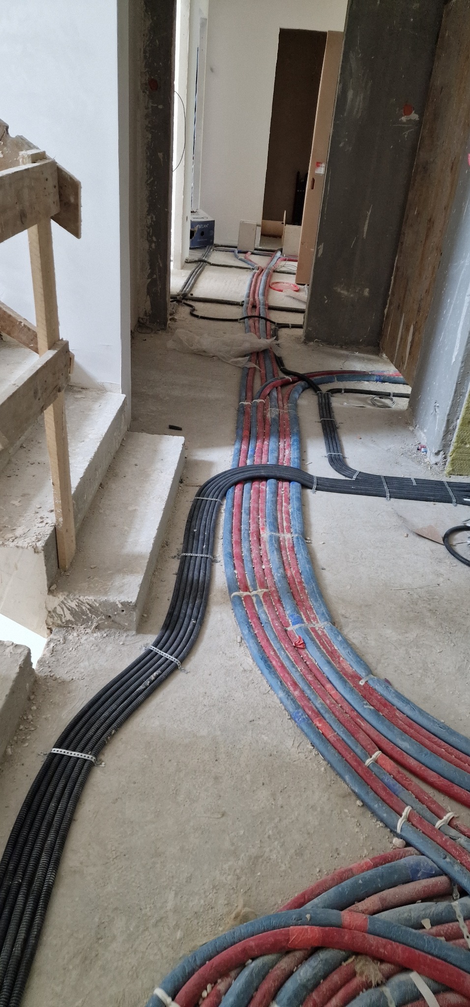 Rzędy czarnych i czerwono-niebieskich rur karbowanych, ułożone na surowym betonowym podłożu w trakcie prac wykończeniowych w budynku, z widocznymi schodami i fragmentami ścian.