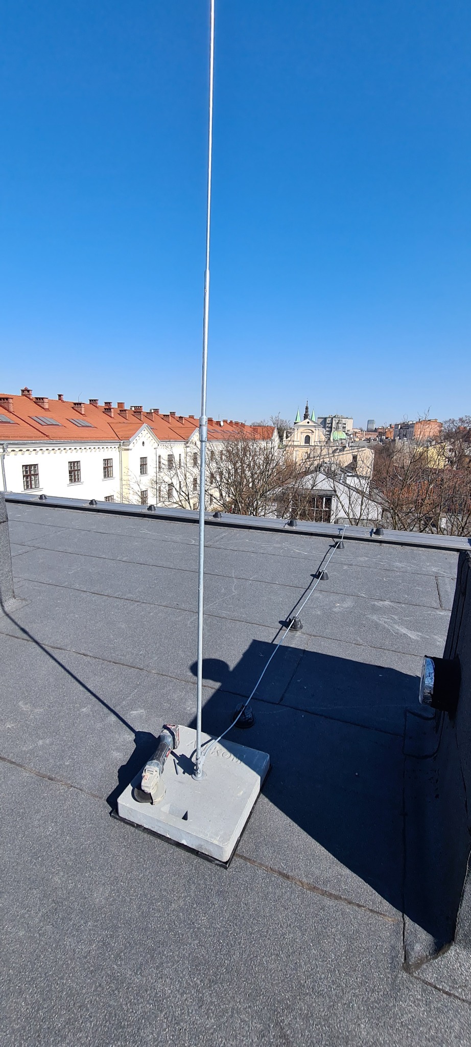 Instalacja odgromowa na dachu budynku z widoczną szlifierką kątową, w tle panorama miasta w słoneczny dzień.
