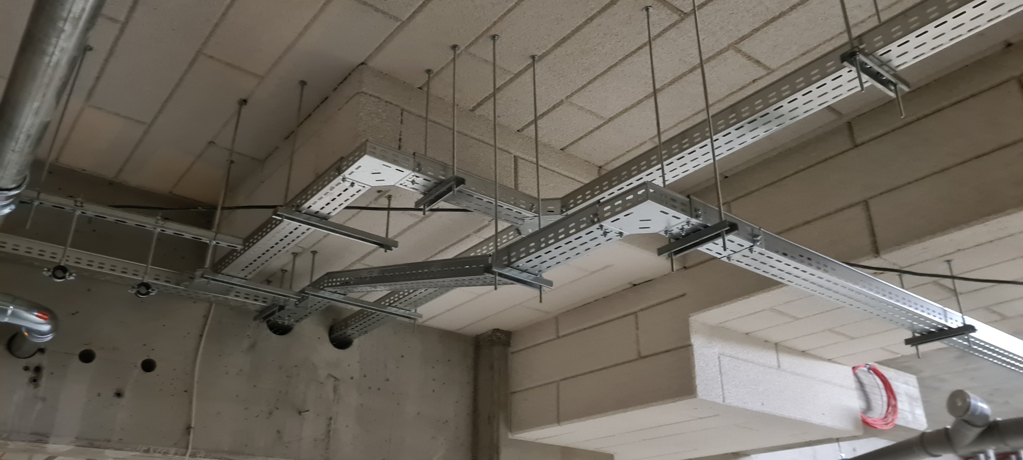 Metalowe koryta kablowe przymocowane do sufitu z bloków betonowych, widoczne przewody i rury instalacyjne.