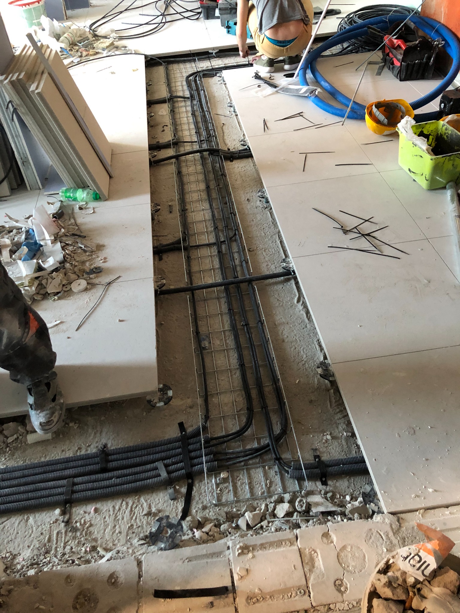 Prace instalacyjne: kable prowadzone w podłodze pod płytkami, widoczne rury karbowane i metalowa siatka, narzędzia i akcesoria na podłodze, osoba w tle.