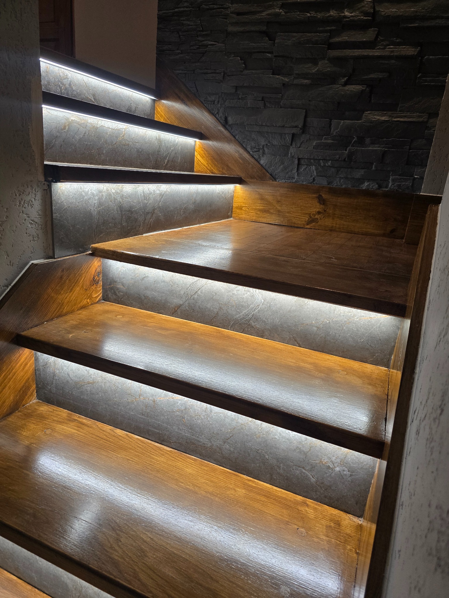 Drewniane schody z podświetleniem LED na każdym stopniu, ściana wykończona kamieniem dekoracyjnym. Ujęcie z dołu, ukazujące detale wykończenia i oświetlenia.