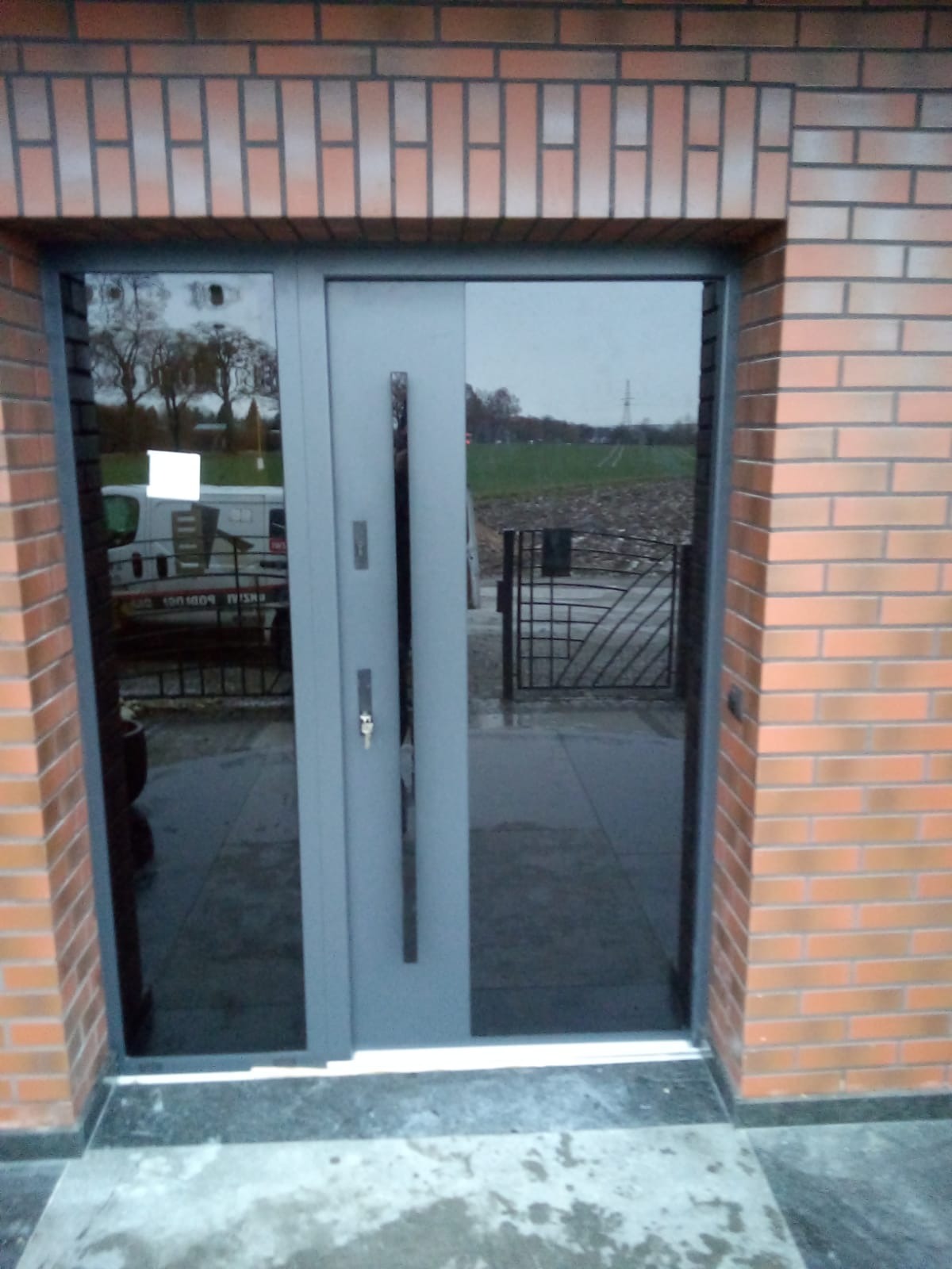 Szare drzwi wejściowe z długim uchwytem i szybą, zamontowane w ścianie z cegły klinkierowej, widok na zewnątrz z odbiciem w szybie. Na zewnątrz widoczny samochód dostawczy i brama.