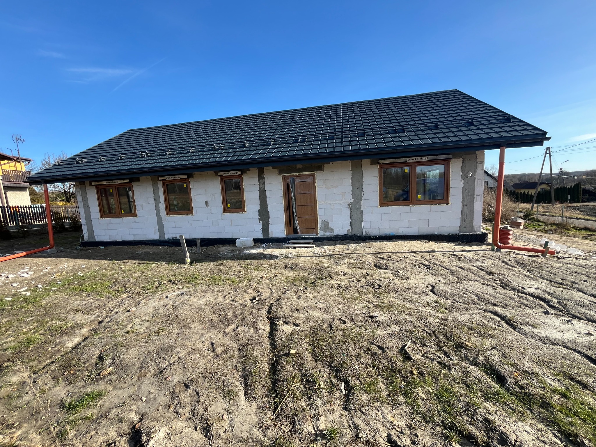 Niedokończony dom jednorodzinny z szarą dachówką i brązowymi ramami okiennymi, widoczne rury spustowe i przygotowany teren wokół budynku.