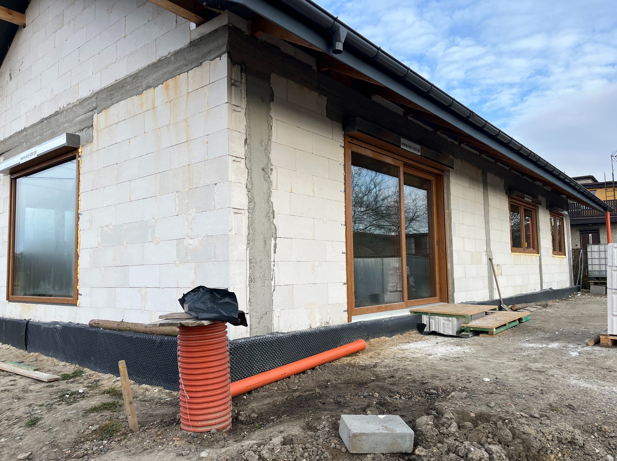 Surowy stan budynku z białych bloczków podczas budowy, widoczne okna z brązowymi ramami, częściowo zamontowane rolety zewnętrzne, izolacja fundamentów, pomarańczowa rura i gruz na placu budowy.