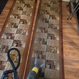 IKB Clean Karolina Bensz - Czyszczenie dywanu za pomocą odkurzacza piorącego z żółtą końcówką na tle dwóch brązowych dywanów w geometryczne wzory.