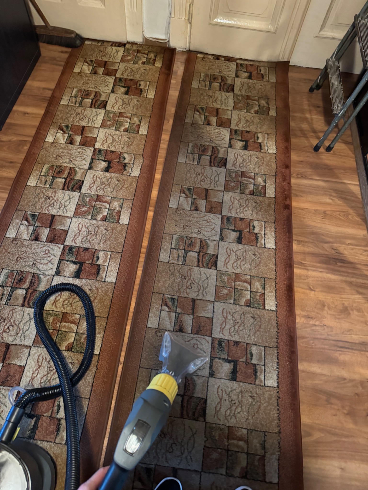 Czyszczenie dywanu za pomocą odkurzacza piorącego z żółtą końcówką na tle dwóch brązowych dywanów w geometryczne wzory.