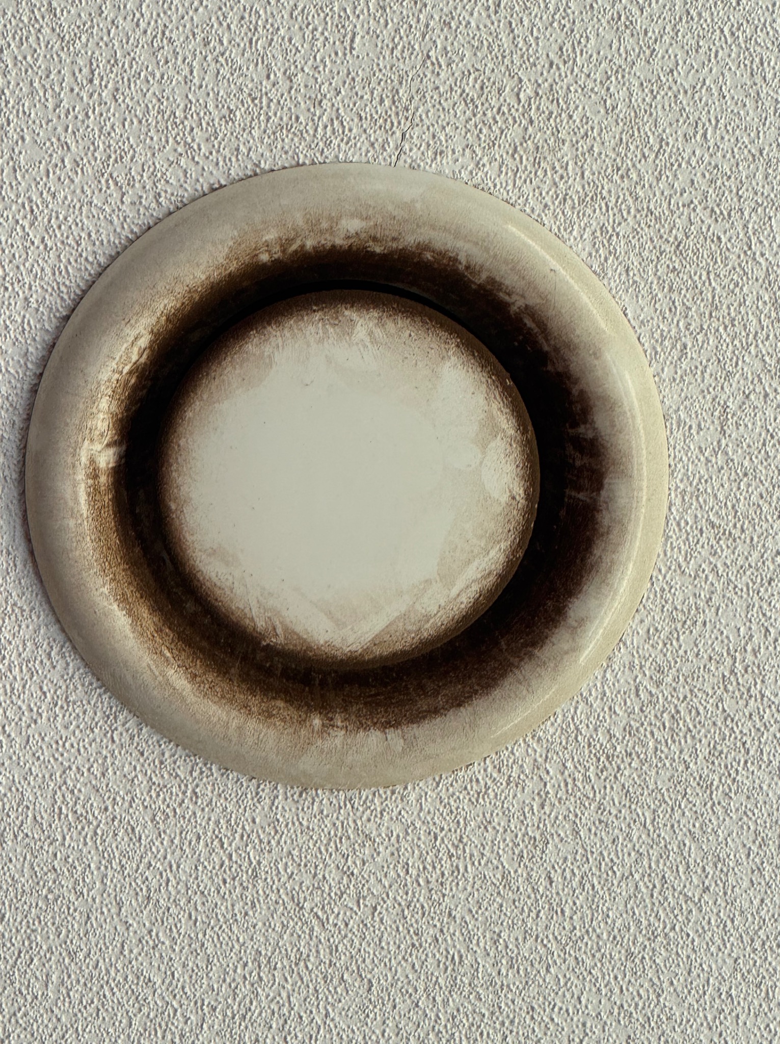 Zabrudzony, okrągły nawiewnik wentylacyjny na teksturowanej, białej ścianie. Widoczne ciemne ślady wokół krawędzi, sugerujące brak czyszczenia.