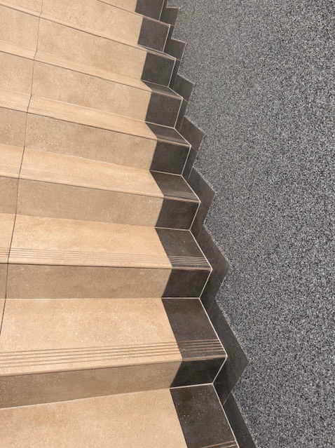 Schody wyłożone beżowymi płytkami z ciemnymi obrzeżami, przylegające do ściany z szarego, drobnego kamienia dekoracyjnego. Widok z góry, ostre światło.