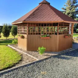 Drewniana altana ogrodowa z brązowym dachem z gontów bitumicznych, ozdobiona wiszącymi doniczkami z kwiatami, otoczona zielonym trawnikiem i ścieżką z kamyków w słoneczny dzień.