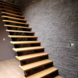 Nowoczesne drewniane schody z czarnymi wspornikami, prowadzące w górę wzdłuż ściany wyłożonej szarym kamieniem dekoracyjnym.