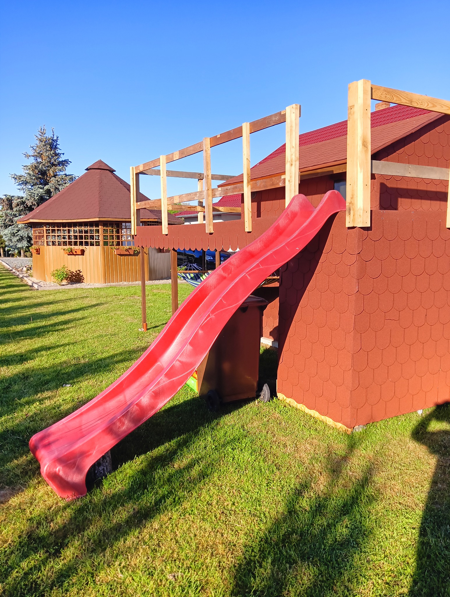 Plac zabaw w trakcie budowy: czerwona zjeżdżalnia, drewniana konstrukcja i altana ogrodowa na trawniku.