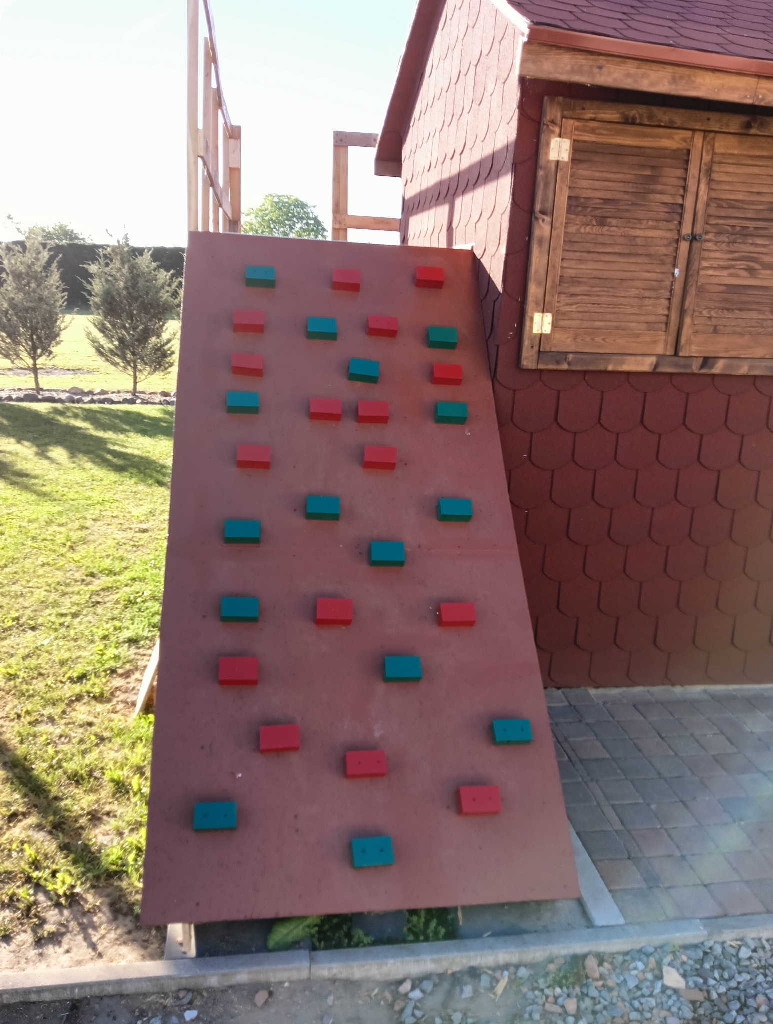 Ściana wspinaczkowa dla dzieci z czerwono-zielonymi uchwytami, przylegająca do drewnianego domku z brązową dachówką, na tle zielonego trawnika i drzew.