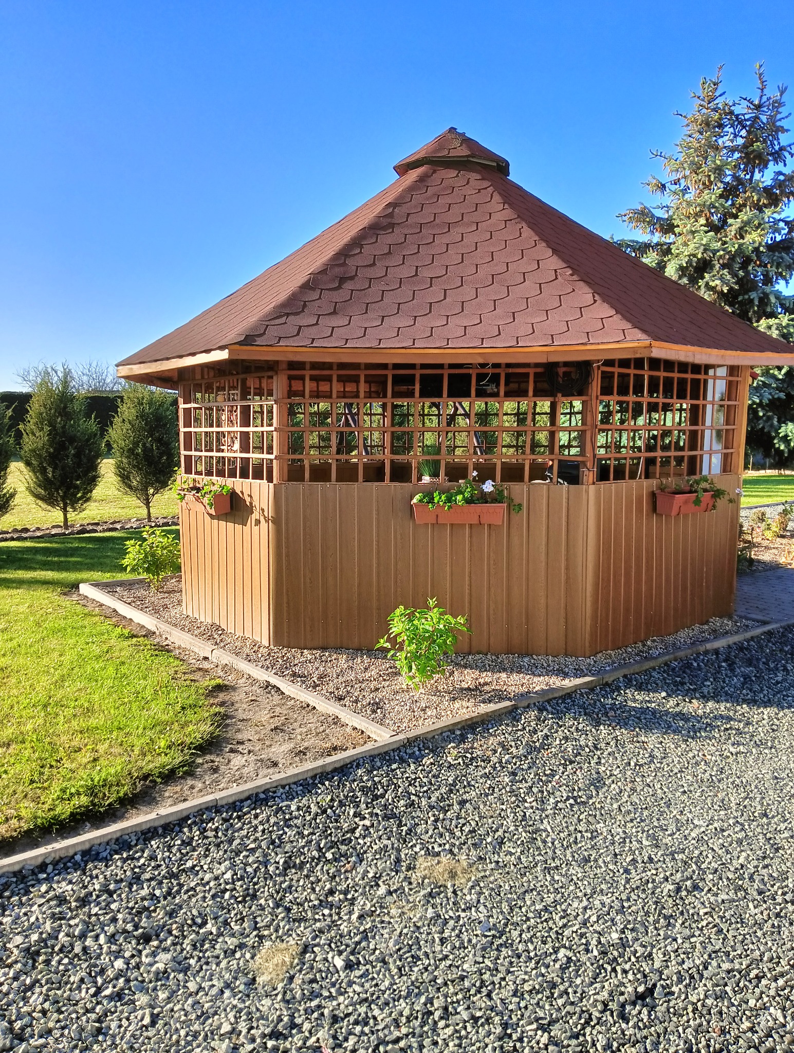 Drewniana altana ogrodowa z brązowym dachem z gontów bitumicznych, ozdobiona wiszącymi doniczkami z kwiatami, otoczona zielonym trawnikiem i ścieżką z kamyków w słoneczny dzień.