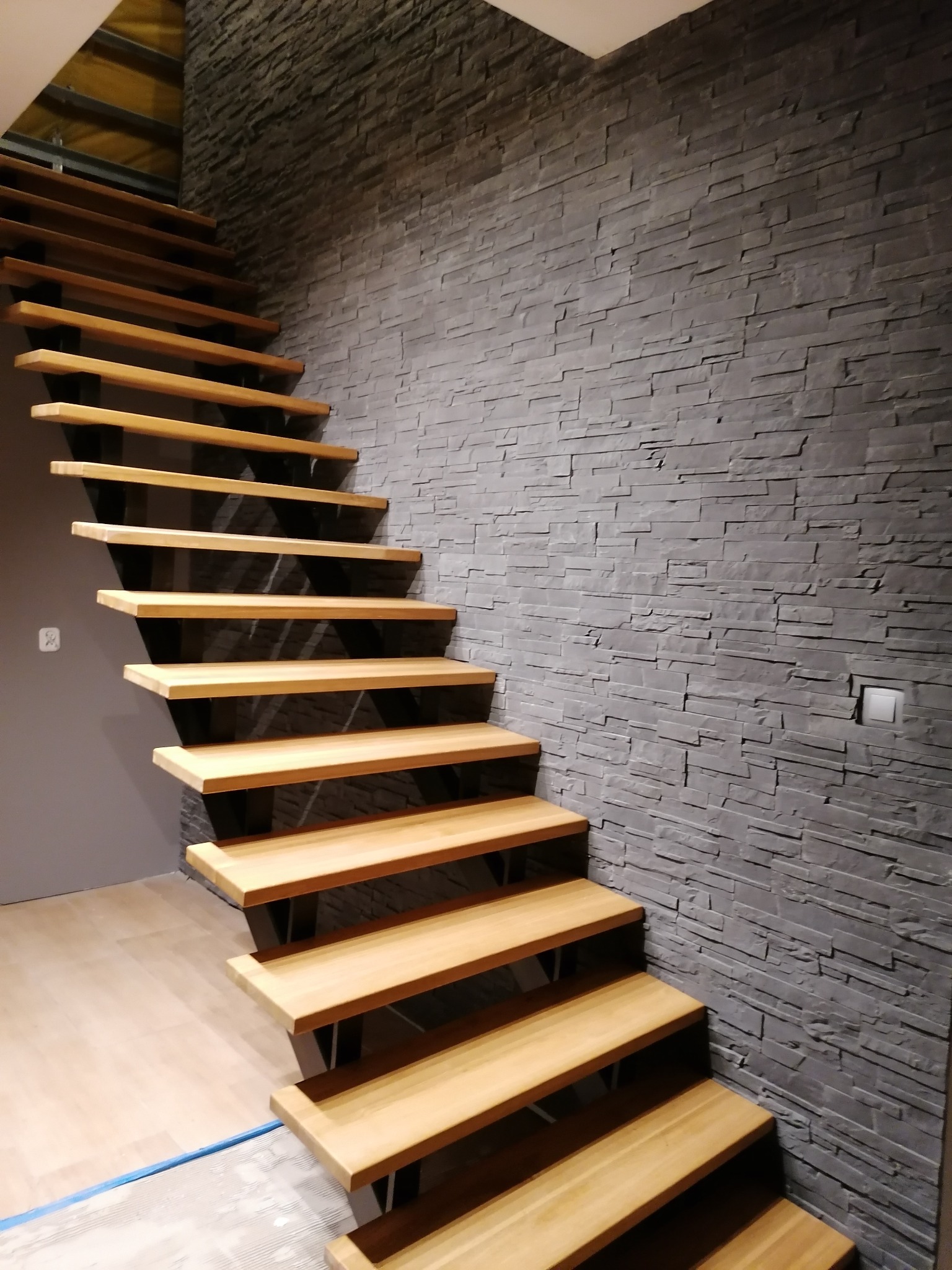 Nowoczesne drewniane schody z czarnymi wspornikami, prowadzące w górę wzdłuż ściany wyłożonej szarym kamieniem dekoracyjnym.