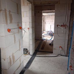 Piotr Wyszyński - Surowy stan instalacji elektrycznej w nowym budynku: pomarańczowe puszki podtynkowe z wystającymi przewodami, kable w peszlach ułożone wzdłuż ściany z betonu komórkowego, widoczne narzędzia...