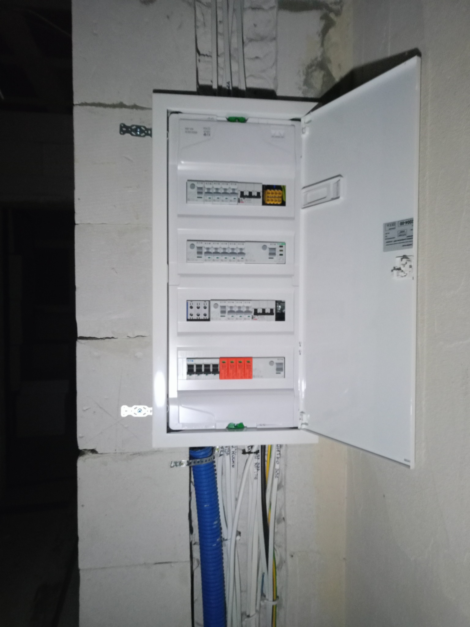 Otwarta skrzynka rozdzielcza z widocznymi bezpiecznikami i przewodami, zamontowana na surowej ścianie z bloczków betonowych.