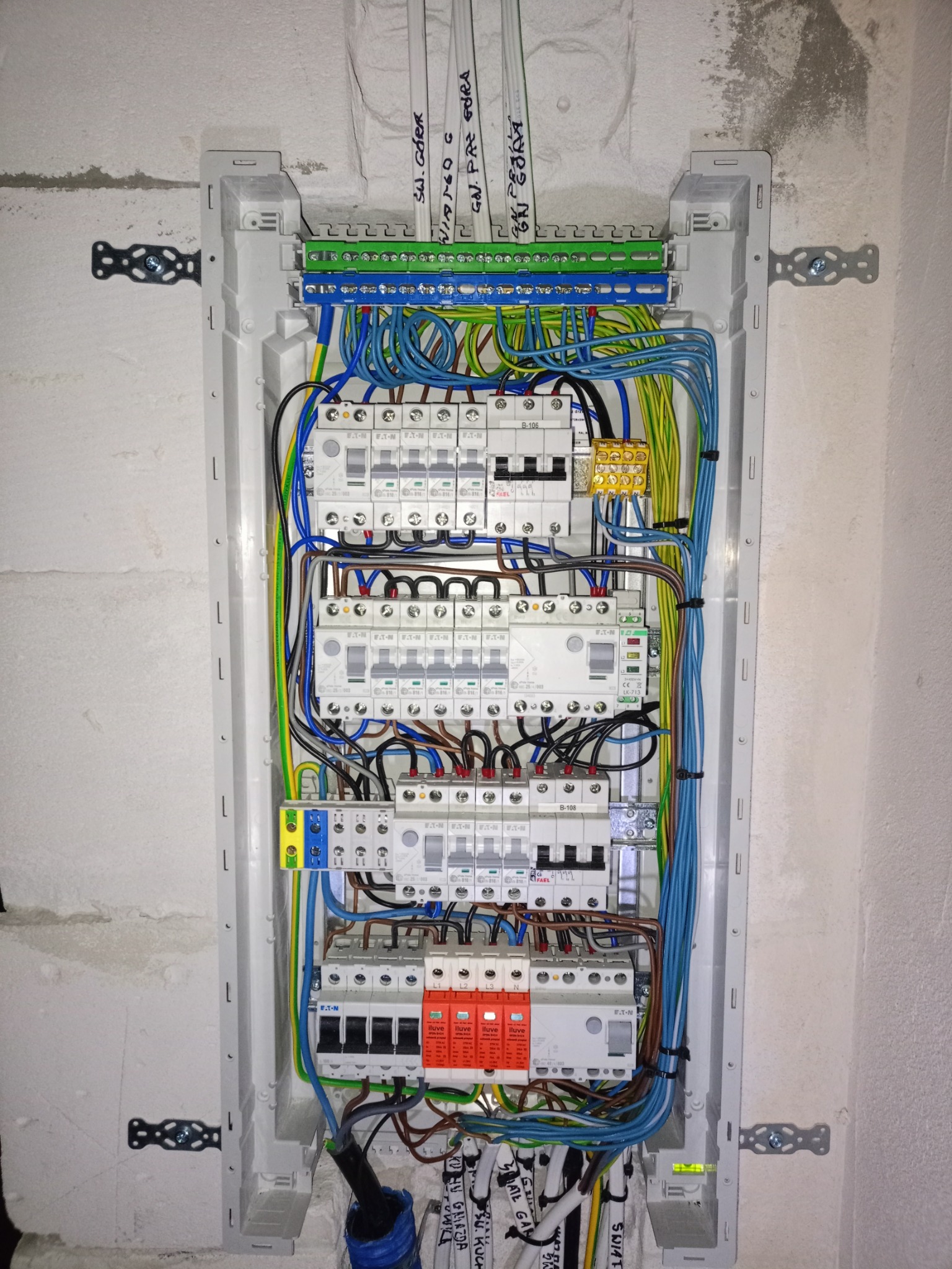 Otwarta skrzynka elektryczna z zamontowanymi bezpiecznikami, zabezpieczeniami przeciwprzepięciowymi i listwami zaciskowymi, widoczne kable w różnych kolorach, zamocowana w surowej ścianie z betonu...