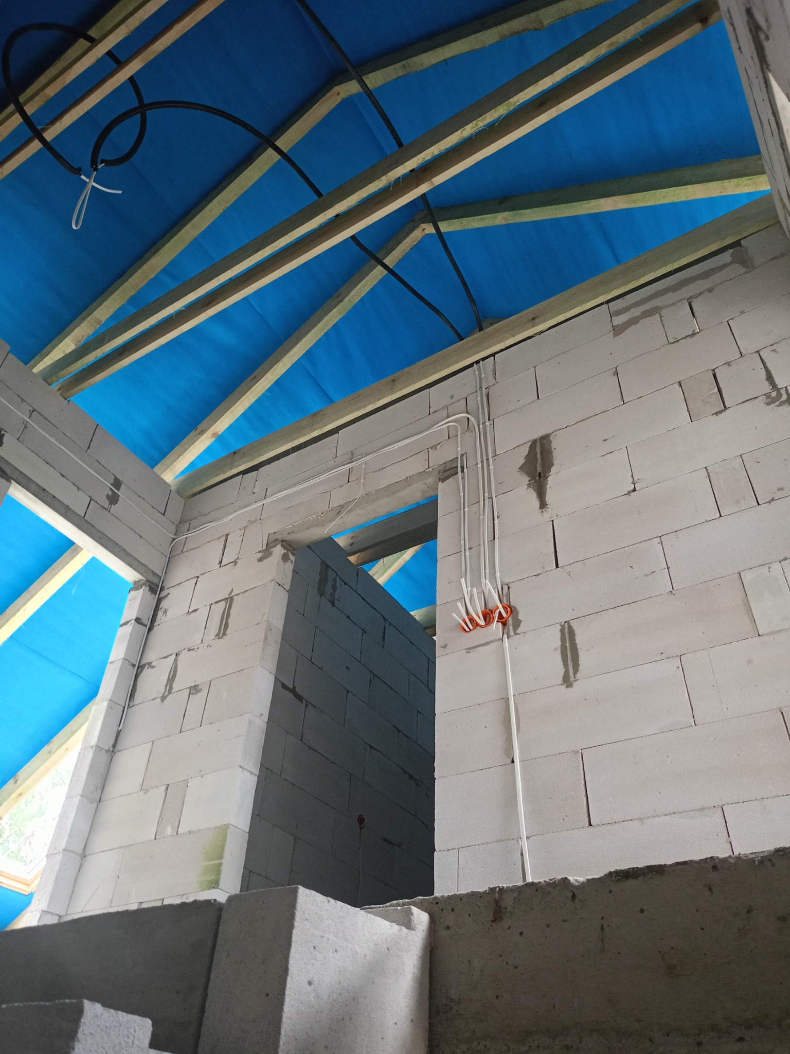 Wnętrze surowego budynku w trakcie budowy, widoczne przewody elektryczne prowadzone po ścianach z białych bloczków, drewniana konstrukcja dachu z niebieską folią zabezpieczającą.