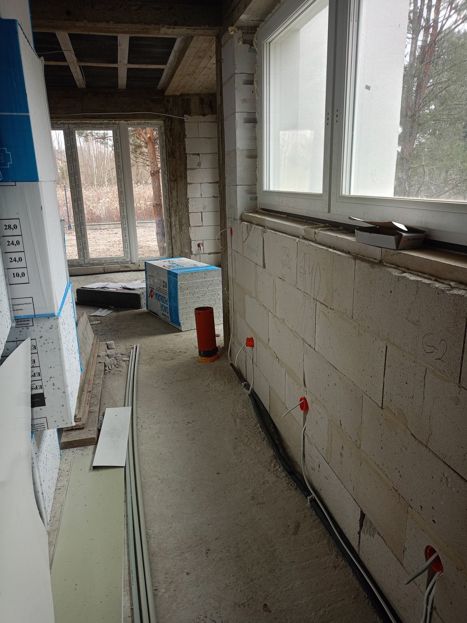 Wnętrze surowego budynku w trakcie prac instalacyjnych; widoczne przewody elektryczne wychodzące z pomarańczowych puszek w ścianie z betonu komórkowego, okno z widokiem na drzewa oraz materiały...