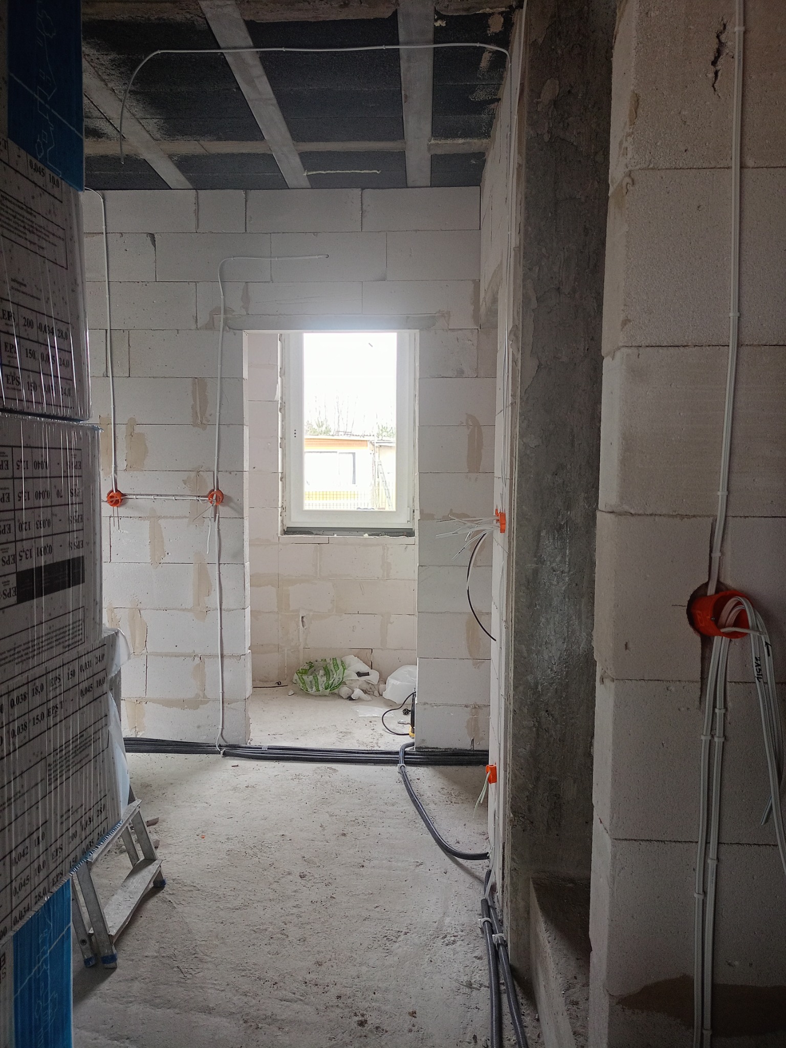 Surowy stan wnętrza w trakcie prac elektrycznych: widoczne kable w peszlach, puszki instalacyjne w ścianach z bloczków, częściowo ułożona instalacja podtynkowa, w tle okno z widokiem na zewnątrz.
