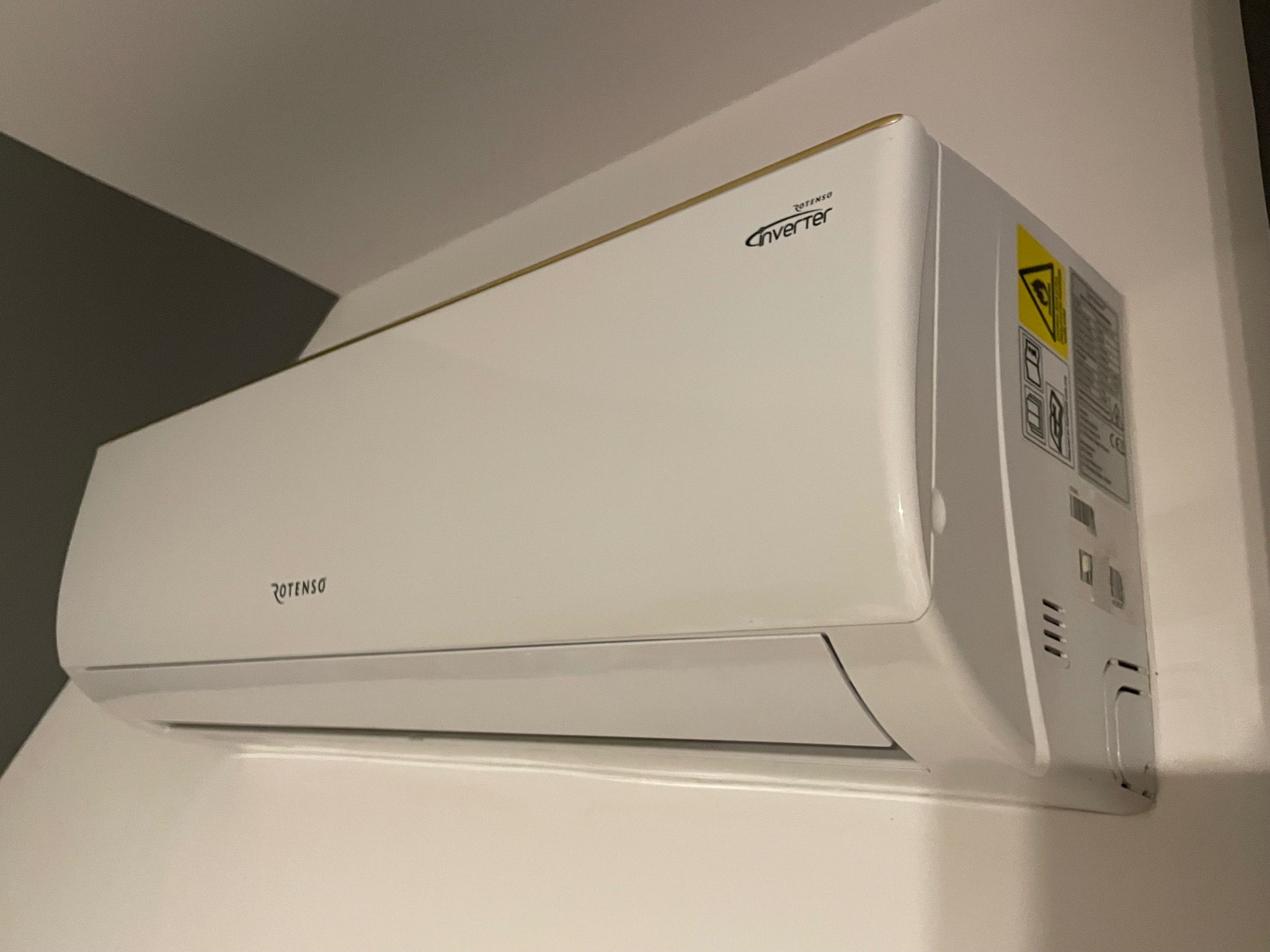 Biała jednostka wewnętrzna klimatyzatora Rotenso Inverter zamontowana na ścianie, widoczna z boku pod kątem, z naklejką ostrzegawczą i specyfikacją techniczną po prawej stronie.
