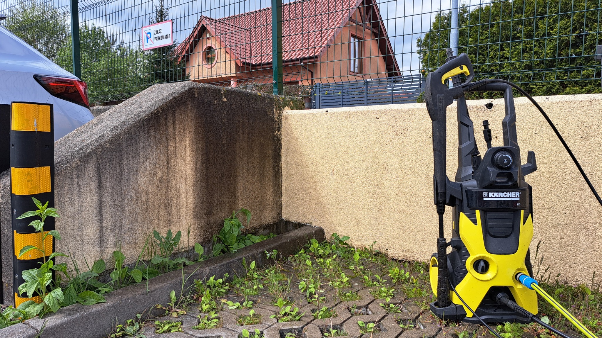Żółto-czarna myjka ciśnieniowa Karcher K5 stoi na betonowej kostce brukowej porośniętej trawą, na tle ściany z widocznymi zabrudzeniami i fragmentem samochodu z żółtymi odblaskami.