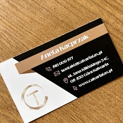 Wizytówka z danymi kontaktowymi Anety Kacprzak, zawierająca numer telefonu, adres e-mail, adres siedziby w Górze Kalwarii i adres strony internetowej, umieszczona na drewnianym biurku.
