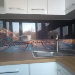 Szklany panel kuchenny z motywem kawy, zamontowany nad zlewem i blatem, widoczne białe szafki.