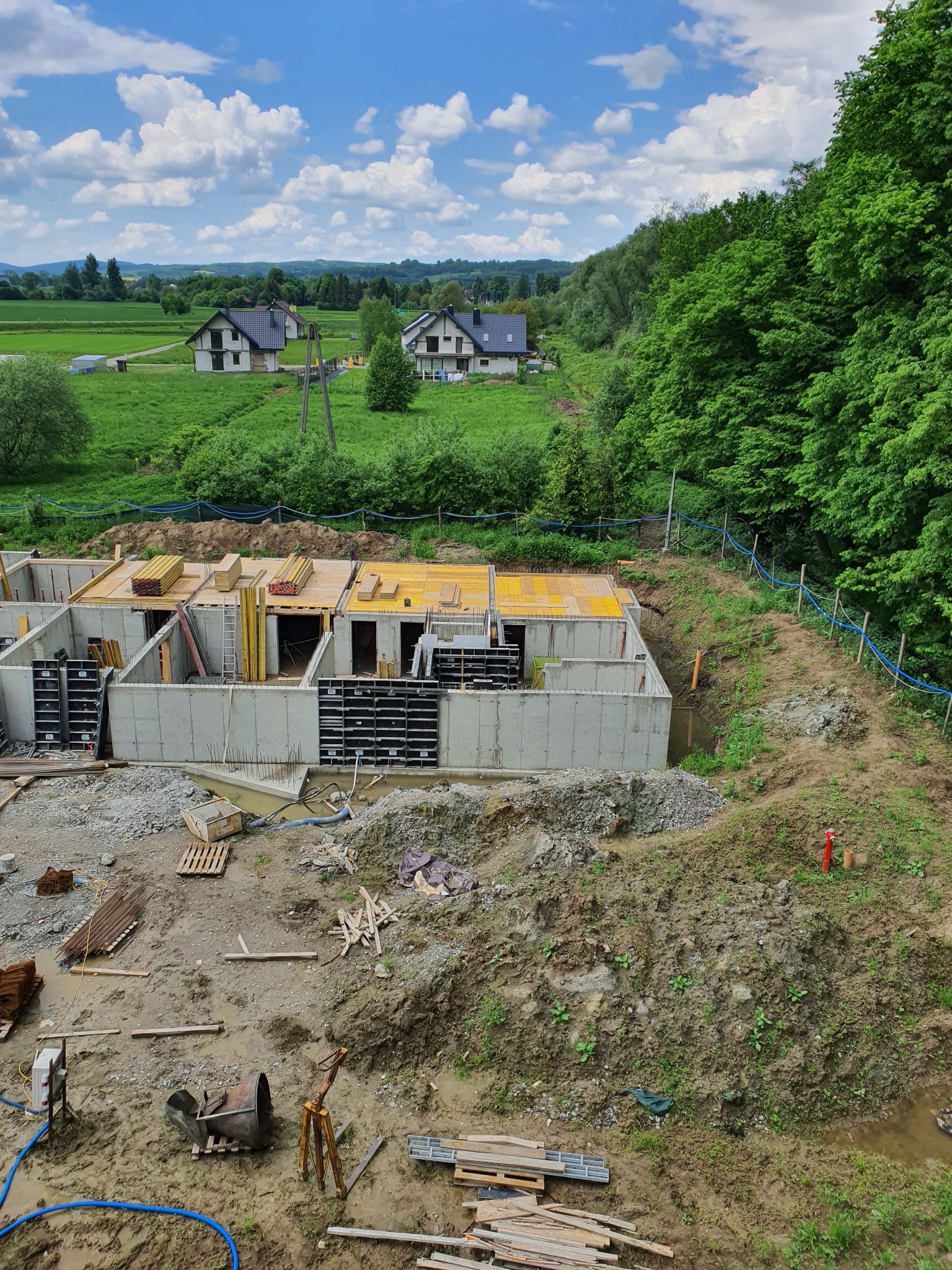 Wysoki kąt widzenia na budowę fundamentów domu jednorodzinnego na tle zielonego krajobrazu z domami w oddali, widoczne szalunki, zbrojenia i drewniane elementy konstrukcyjne.