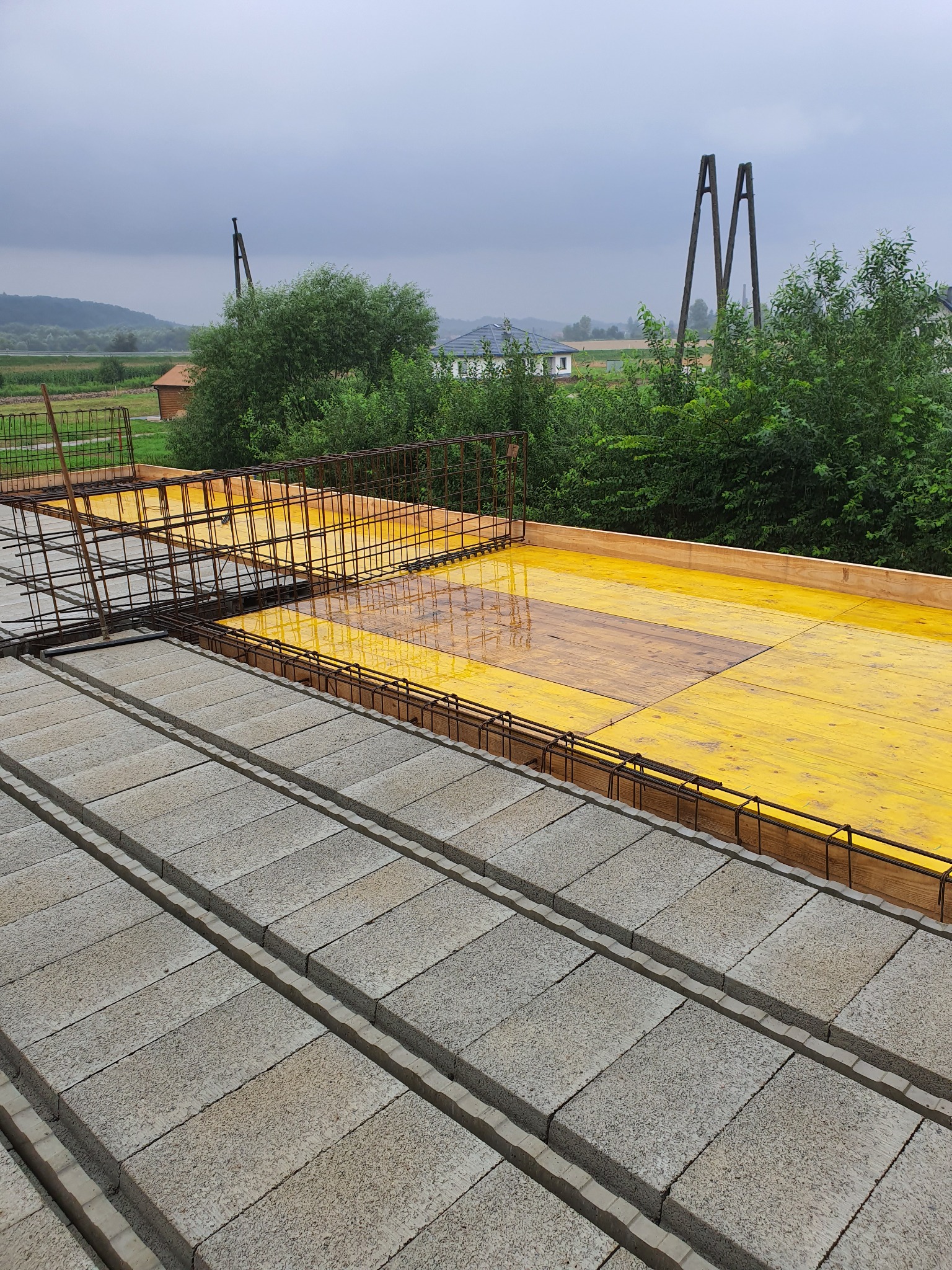 Szare bloczki betonowe ułożone rzędami z widocznym zbrojeniem i żółtą wodoodporną membraną, przygotowane do zalania betonem, w tle krajobraz wiejski w pochmurny dzień.
