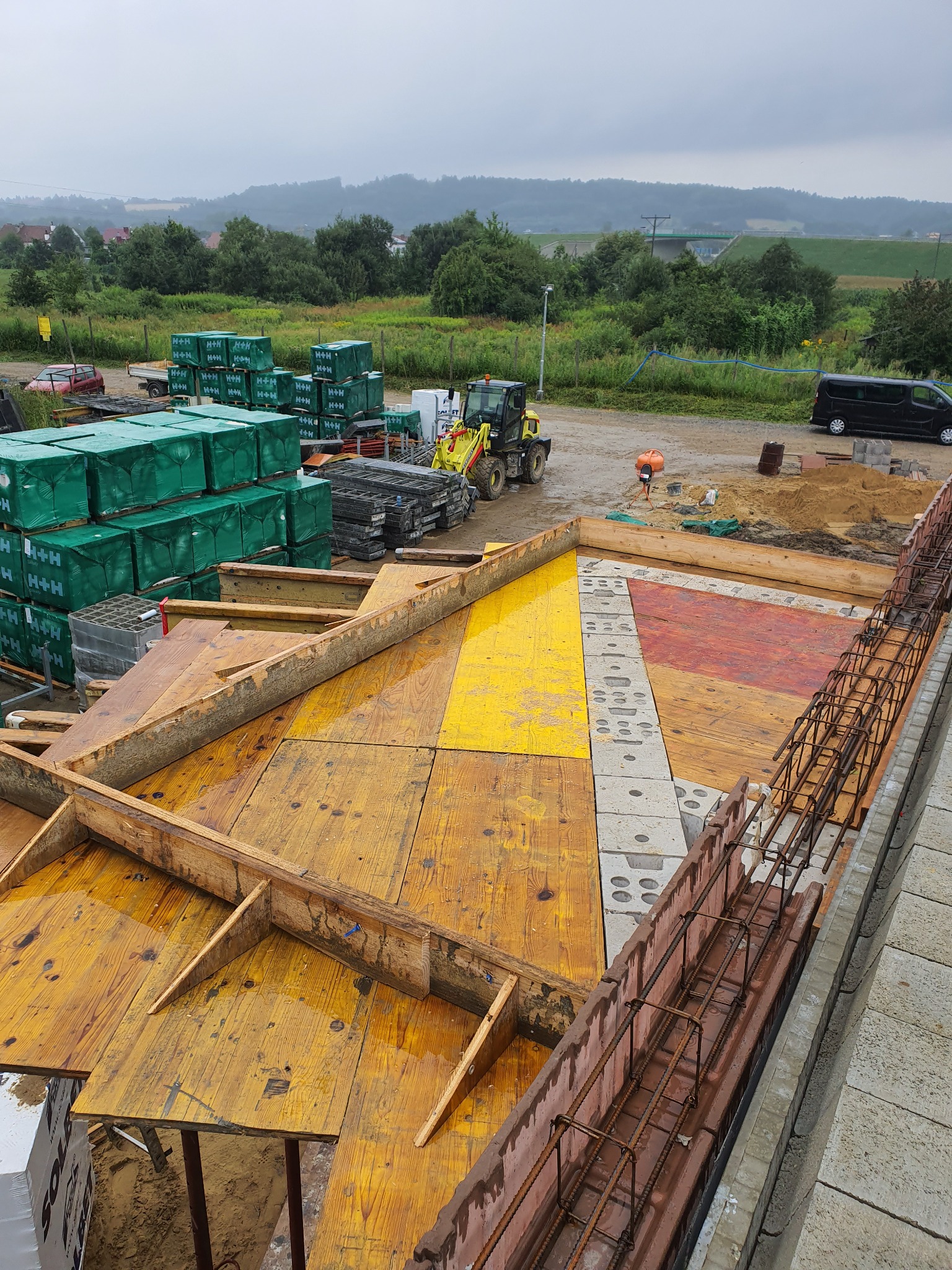 Widok z góry na plac budowy z deskowaniem i zbrojeniem przygotowanym pod wylewkę fundamentów, w tle zaparkowany samochód dostawczy i koparka.