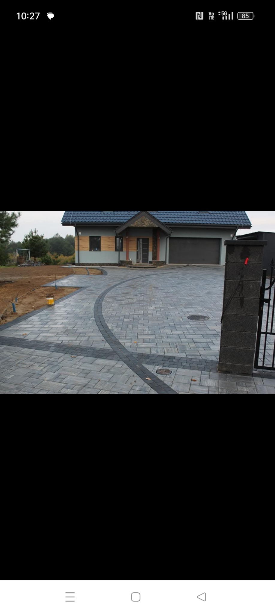 Ułożona szara kostka brukowa z ciemniejszym obramowaniem prowadząca do domu z garażem, widoczny fragment niezakończonych prac ziemnych po lewej stronie.
