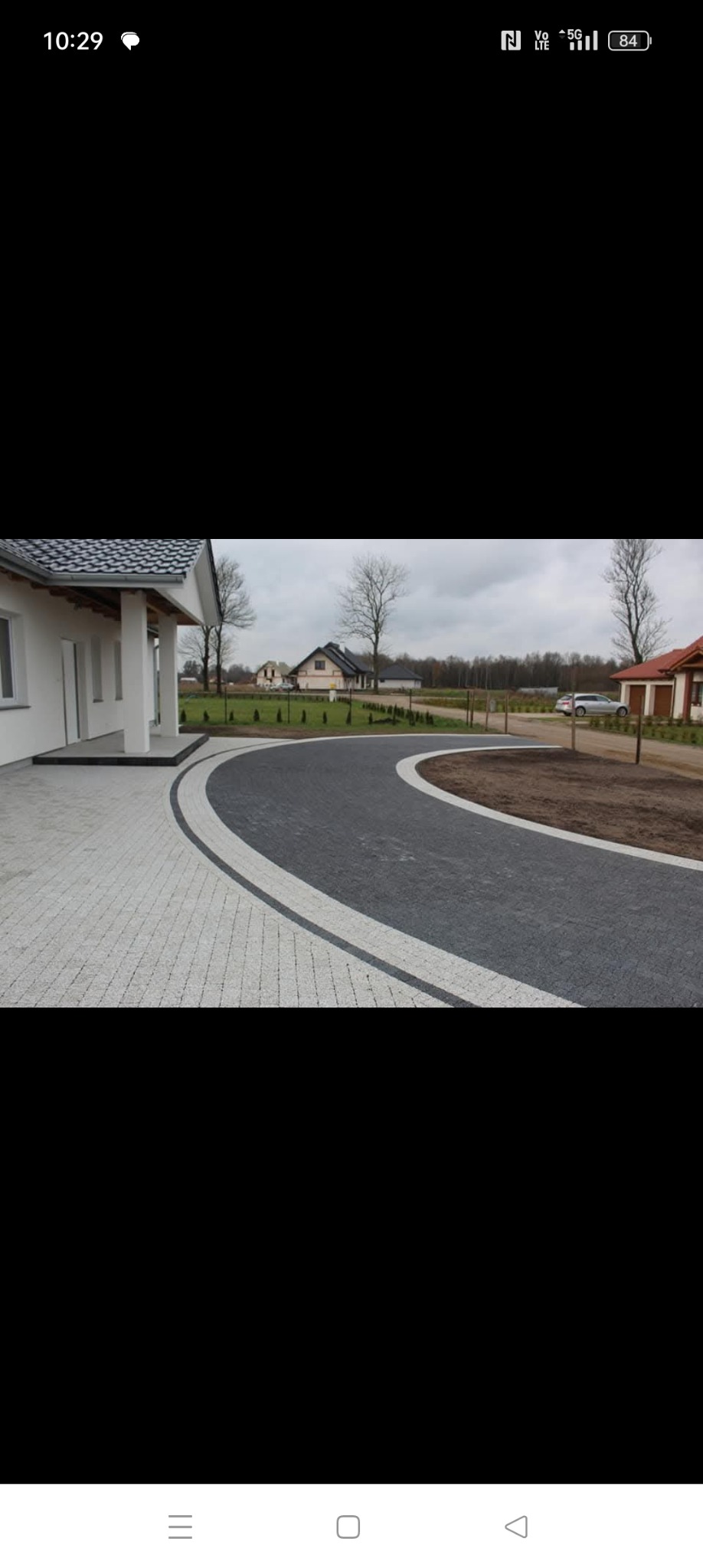 Ułożona jasnoszara kostka brukowa przed domem, z ciemnoszarą kostką tworzącą zakrzywiony podjazd obrzeżony pasem jasnej kostki, w tle widoczne domy i nagie drzewa.