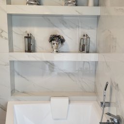 USŁUGI OGÓLNOBUDOWLANE PIOTR PŁÓCIENNIK - Biała wanna z hydromasażem pod ścianą z marmurowych płytek. Na dwóch półkach dekoracje: srebrne doniczki z zielonymi roślinami, lampiony i popiersie z kwiatami.