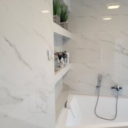 USŁUGI OGÓLNOBUDOWLANE PIOTR PŁÓCIENNIK - Biała łazienka wykończona płytkami imitującymi marmur z wnękami na dekoracje, widoczna wanna z chromowaną armaturą i słuchawką prysznicową.