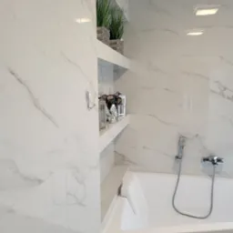 Biała łazienka wykończona płytkami imitującymi marmur z wnękami na dekoracje, widoczna wanna z chromowaną armaturą i słuchawką prysznicową.