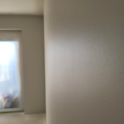 USŁUGI OGÓLNOBUDOWLANE PIOTR PŁÓCIENNIK - Świeżo pomalowana ściana w jasnym odcieniu beżu, widoczne delikatne smugi pędzla, w tle okno z zasłoniętą szybą, częściowo widoczna podłoga.