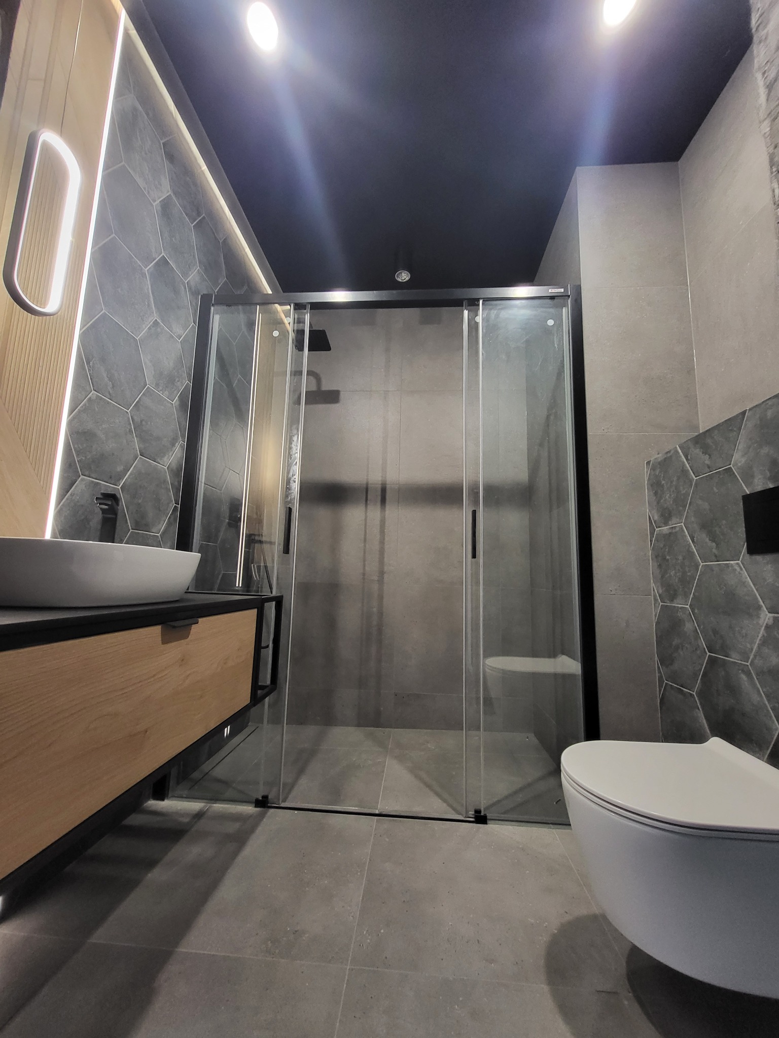 Nowoczesna łazienka w odcieniach szarości z heksagonalnymi płytkami na ścianie, drewnianą szafką pod umywalką z czarnymi elementami i kabiną prysznicową z czarną ramą.