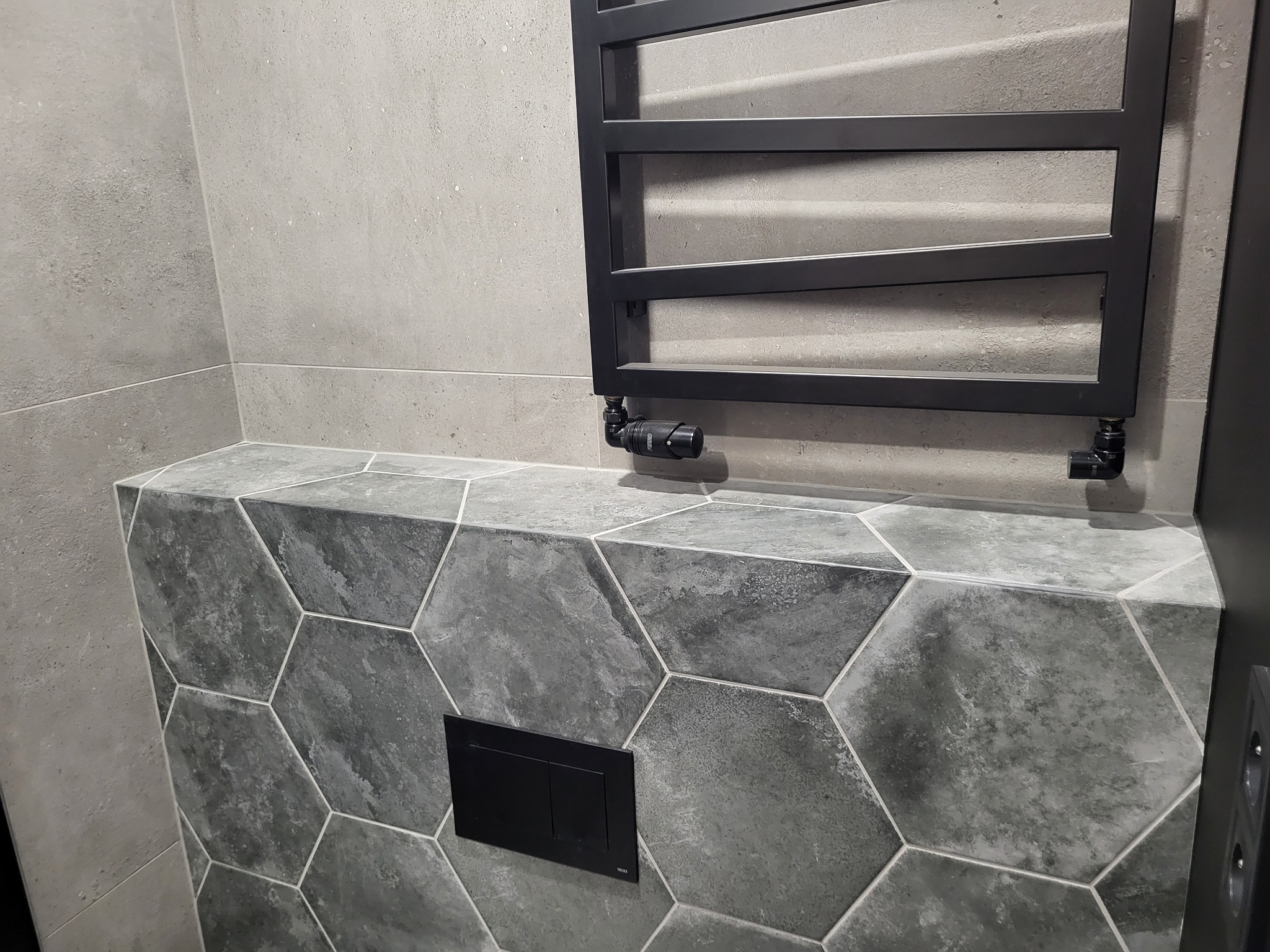 Szara łazienka z heksagonalnymi płytkami na zabudowie stelaża WC, grzejnik drabinkowy w kolorze czarnym, szare płytki ścienne.