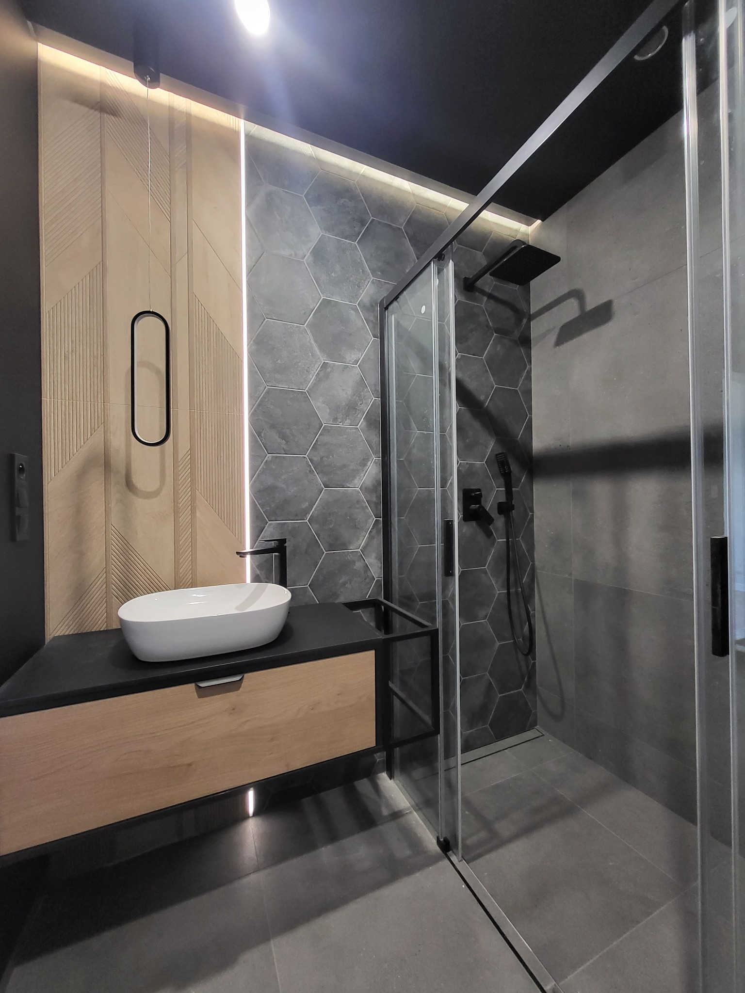 Nowoczesna łazienka z kabiną prysznicową typu walk-in, umywalką nablatową na szafce z drewnianym frontem, heksagonalnymi płytkami na ścianie prysznica i ozdobną ścianą z drewnopodobnych paneli.