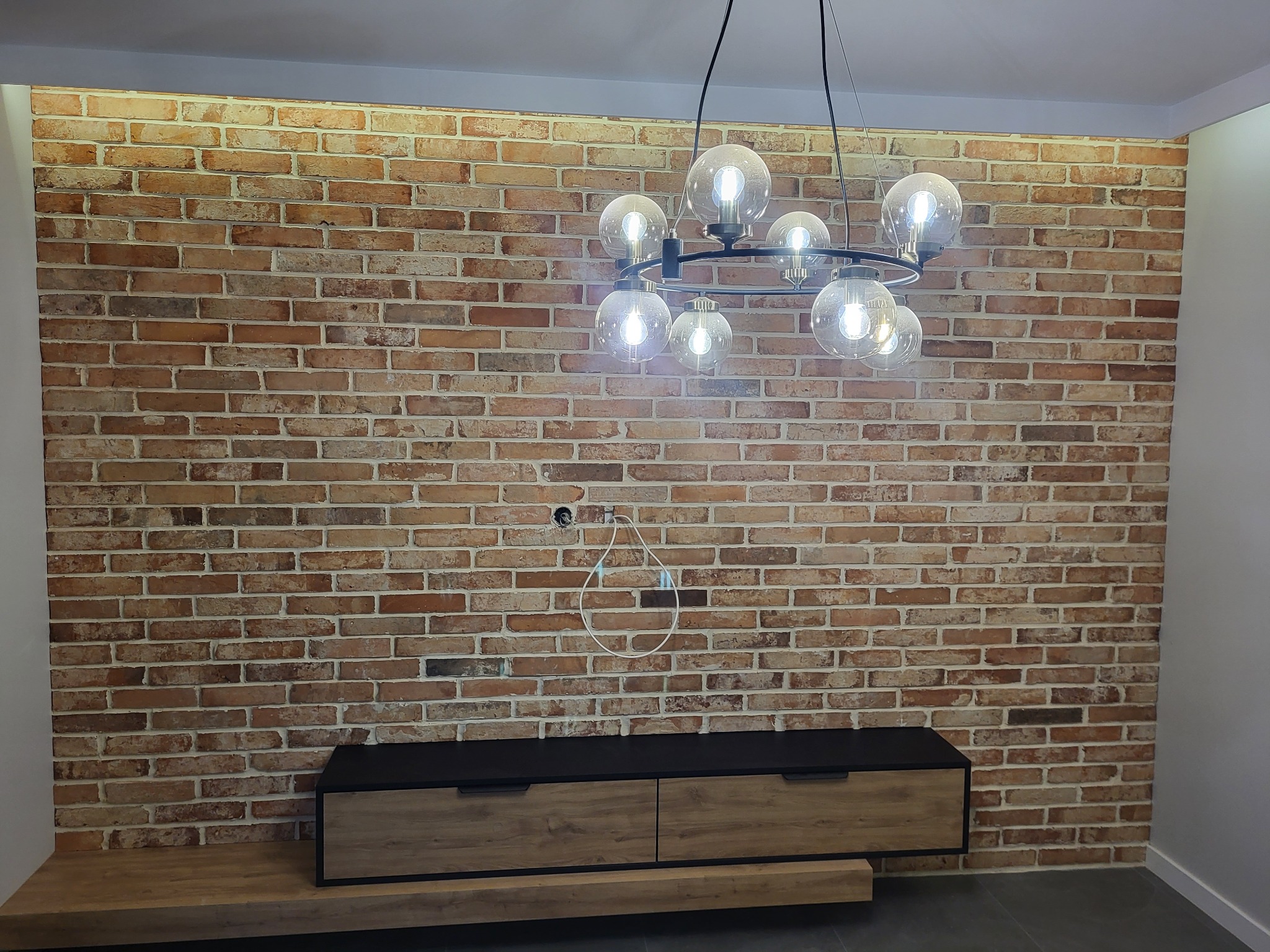 Ściana z cegły klinkierowej z widocznym otworem na kabel i wiszącym przewodem, podświetlona przez nowoczesną lampę z kulistymi kloszami, z szafką RTV na tle ściany.