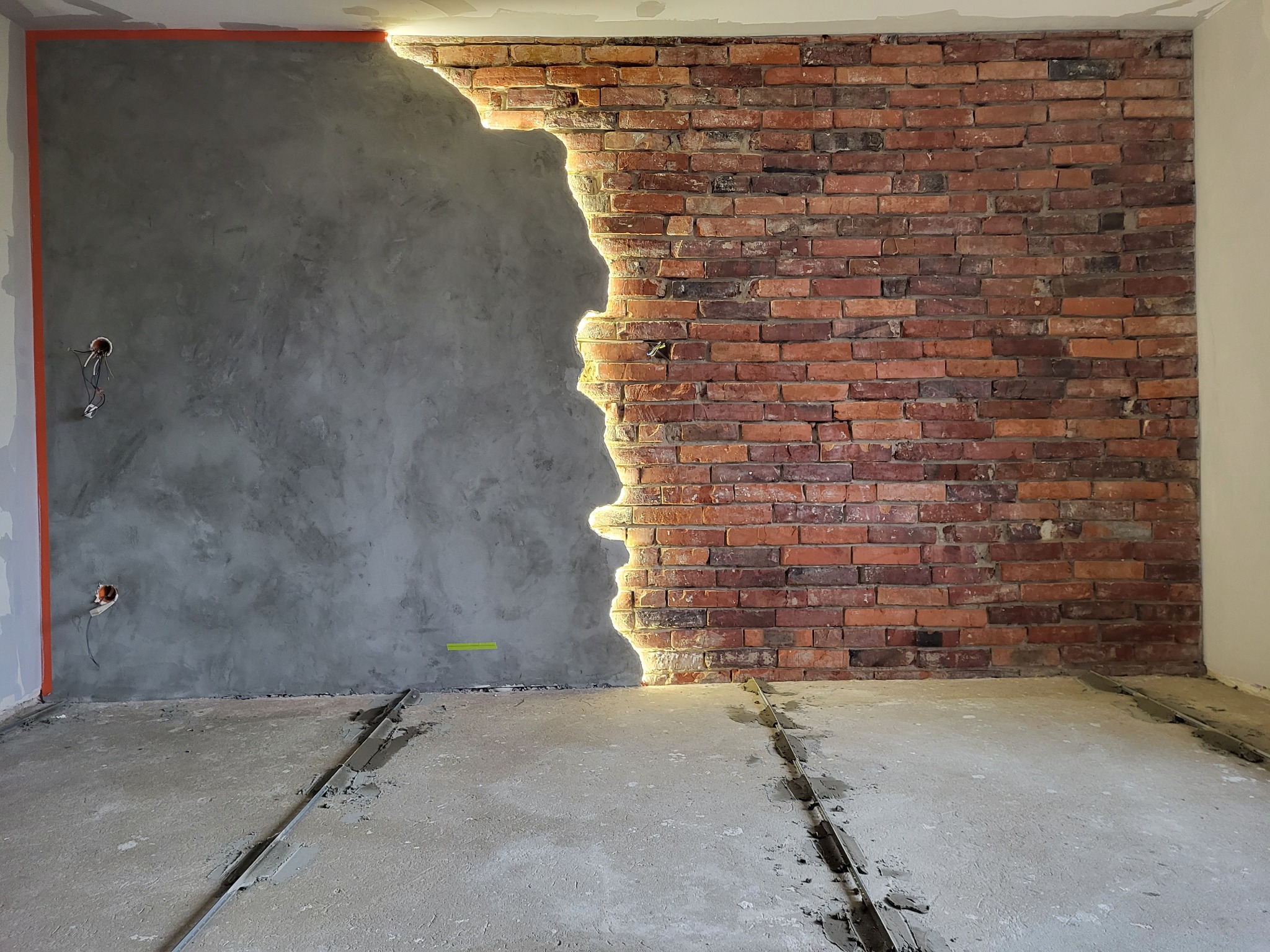 Ściana w trakcie remontu: połączenie surowej cegły i szarego tynku strukturalnego, z widocznymi przewodami elektrycznymi i elementami przygotowania podłoża na podłodze.