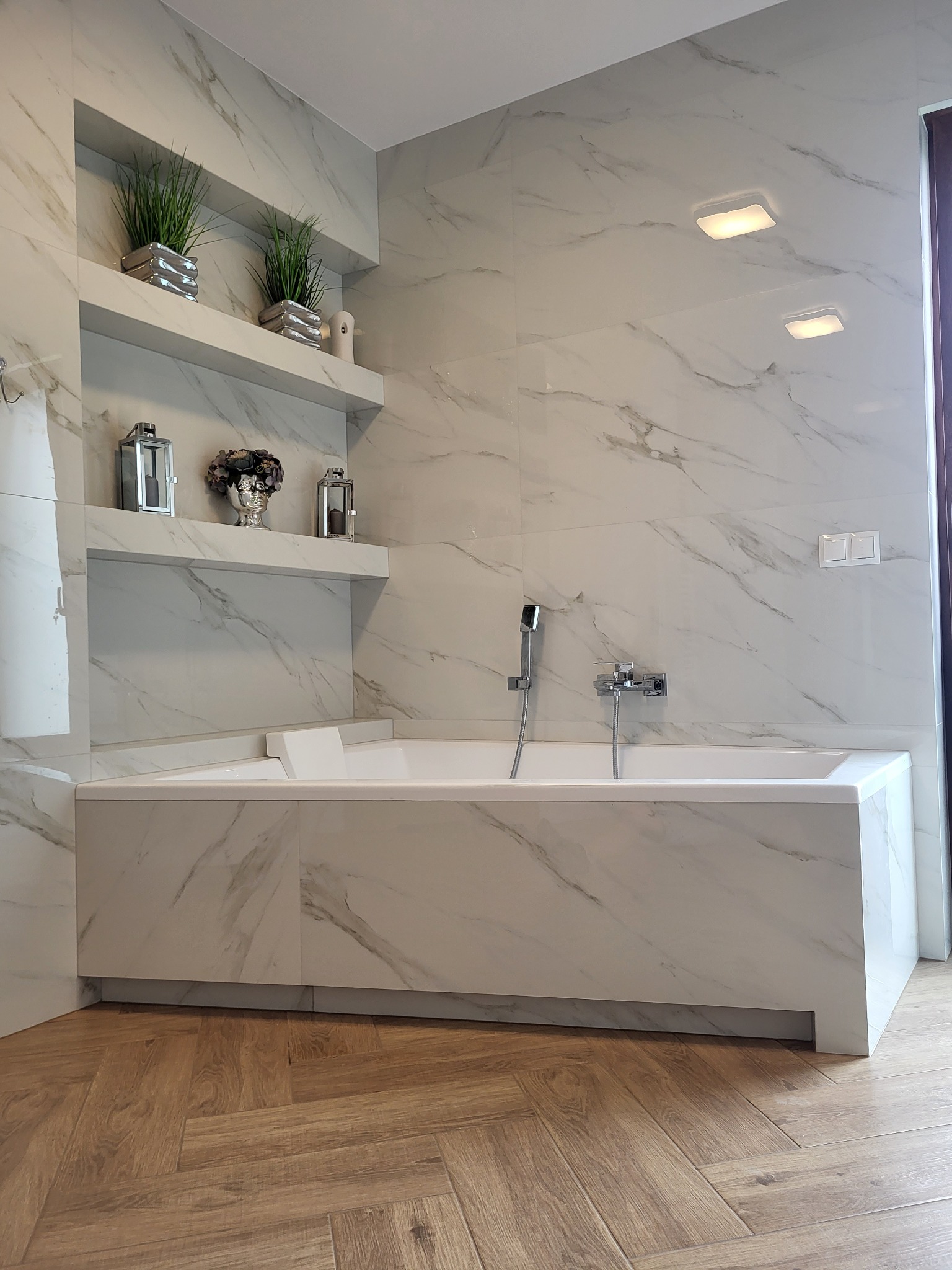 Nowoczesna łazienka wykończona białymi płytkami imitującymi marmur, z wanną obudowaną tym samym materiałem i drewnianą podłogą ułożoną w jodełkę; na półkach dekoracyjne lampiony i rośliny.