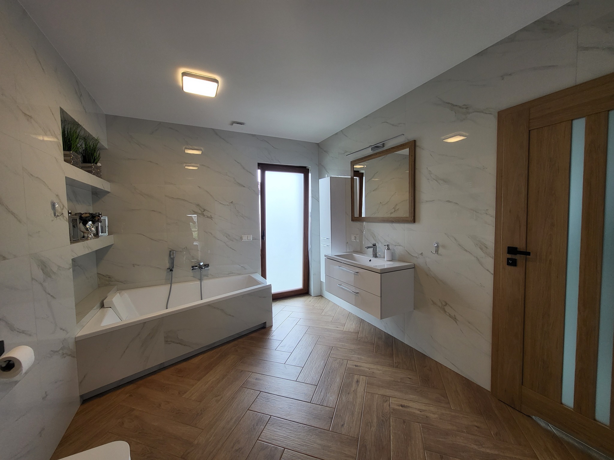 Jasna łazienka z marmurowymi płytkami, drewnianą podłogą ułożoną w jodełkę, białą wanną narożną, wiszącą szafką z umywalką i drewnianymi drzwiami z przeszkleniami.