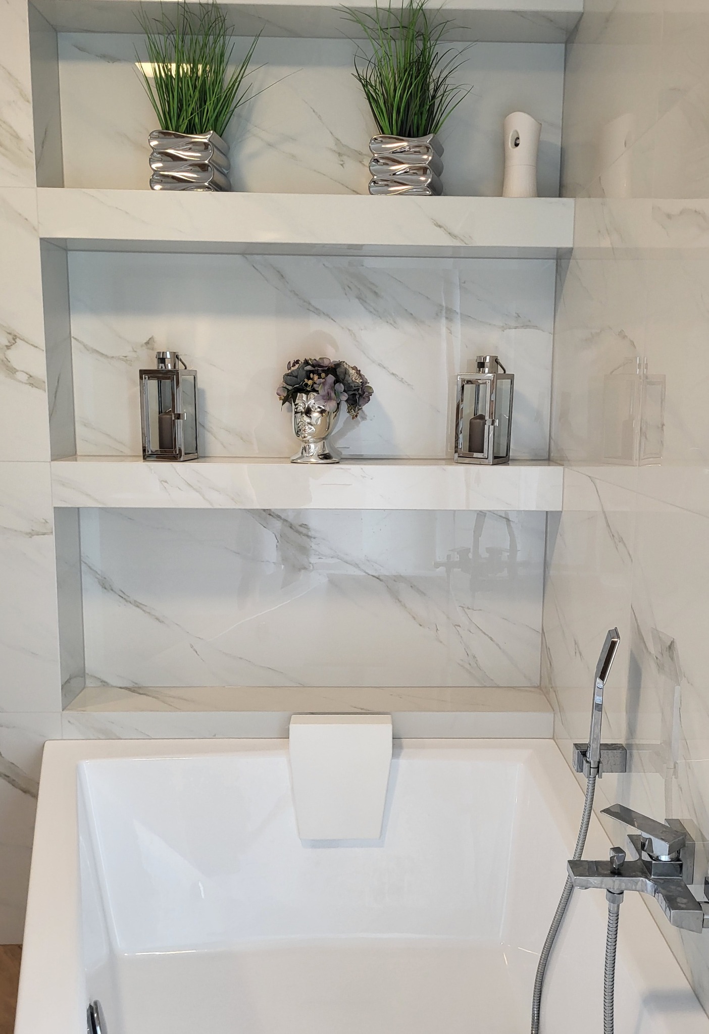 Biała wanna z hydromasażem pod ścianą z marmurowych płytek. Na dwóch półkach dekoracje: srebrne doniczki z zielonymi roślinami, lampiony i popiersie z kwiatami.