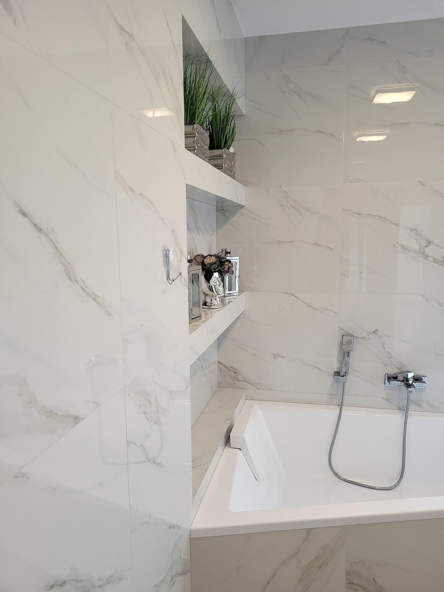 Biała łazienka wykończona płytkami imitującymi marmur z wnękami na dekoracje, widoczna wanna z chromowaną armaturą i słuchawką prysznicową.