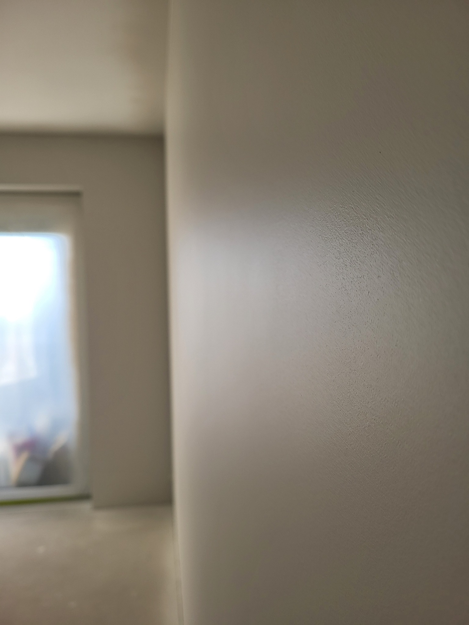 Świeżo pomalowana ściana w jasnym odcieniu beżu, widoczne delikatne smugi pędzla, w tle okno z zasłoniętą szybą, częściowo widoczna podłoga.