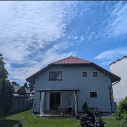 Grzegorz Zabrzeński EKOPRO - Dom z elewacją w odcieniach błękitu, z czerwoną dachówką. Przy elewacji stoi drabina i myjka ciśnieniowa. Widoczne niebo z chmurami i trawnik.