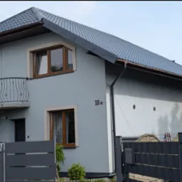 Dom jednorodzinny z szarą elewacją i ciemnym dachem z blachodachówki, widoczne rynny, okna z brązowymi ramami i balkon z ozdobną balustradą. Numer domu 15B.